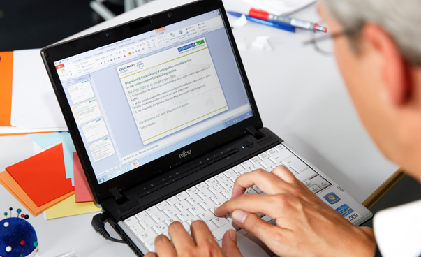 Eine Person ist ausschnitthaft von hinten zu sehen; beide Hände liegen auf der Tastatur eines Laptops. Auf dem Bildschirm sind schemenhaft die Logos von Engagament Global und der Servicestelle zu sehen.