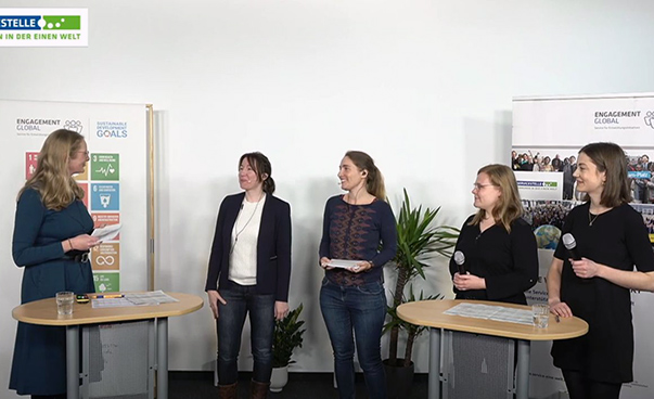 Die Moderatorin spricht vier Frauen an, die Hochtischen stehen. Im Hintergrund sind Aufsteller mit dem Engagement Global-Logo sowie den SDG-Symbolen zu sehen.