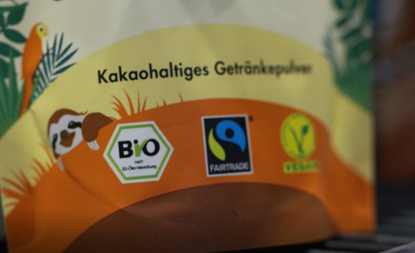 Ein Teil einer Verpackung eines kakaohaltigen Getränkepulvers ist zu sehen; Darauf sind Dschungeltiere und Pfalnzen sowie die Bio-, Fairtrade und Vegansymbole zu sehen.