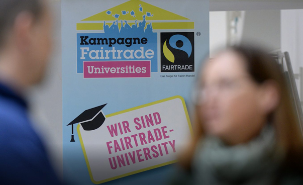 Unscharf im Vordergrund zwei Personen; dahinter ein Fairtrade-Plakat mit der Aufschrift: "Wir sind Fairtrade-University"