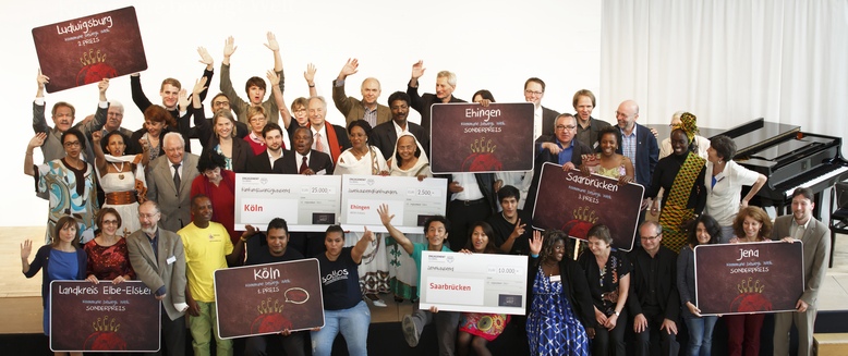Alle Preisträgerinnen und Preisträger des Wettbewerbs Kommune bewegt Welt 2014 stehen auf einer Bühne und halten Schilder mit den Namen der Gewinnerstädte in den Händen.