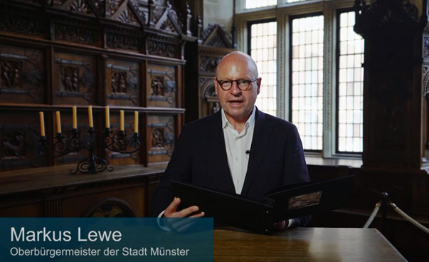 Der Oberbürgermeister von Münster, Markus Lewe, spricht in einem historischen Saal in die Kamera.