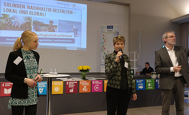 Zwei Frauen und ein Mann stehen vor einer Leinwand, auf der steht: Solingen nachhaltig gestalten - global und lokal. Die Frau in der Mitte spricht in ein Mikrofon.
