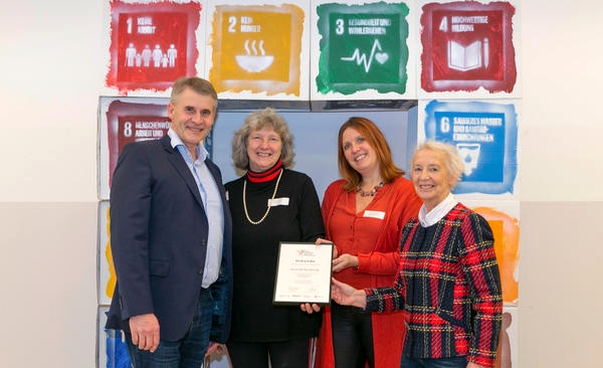 Ein Mann und drei Frauen stehen mit einer Urkunde vor den Symbolen der globalen Nachhaltigkeitsziele
