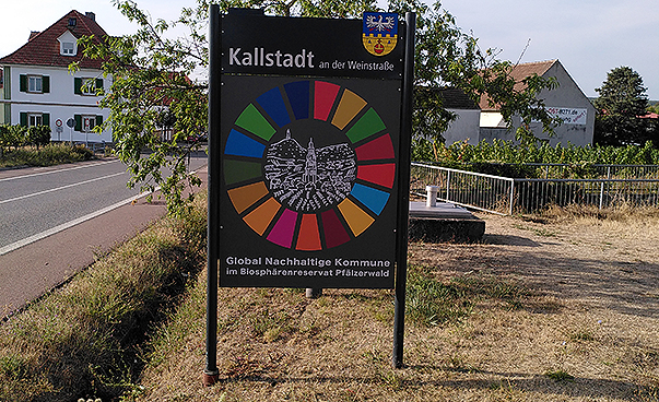 Auf einem großen Schild befindet sich in einem SDG-Kreis die Silhouette von Kallstadt an der Weinstraße, darunter steht „Global Nachhaltige Kommune im Biosphärenreservat Pfälzerwald“