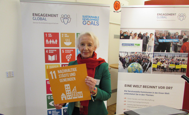 Frau Turmann hält ein Plakat mit dem globalen Nachhaltigkeitsziel 11 vor sich; im Hintergrund sind Plakate von EG und SKEW zu sehen.