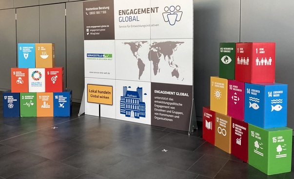 17 Würfel mit den Symbolen der Globalen Nachhaltigkeitsziele stehen links und rechts von einer Stellwand, das das Angebot der Servicestelle Kommunen in der Einen Welt zusammenfasst.