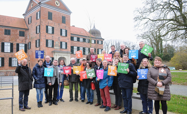 EIne Gruppe lächelt in Richung der Kamera, einige Personen halten Plakate mit den globalen Nachhaltigkeitssymbolen in die Höhe.