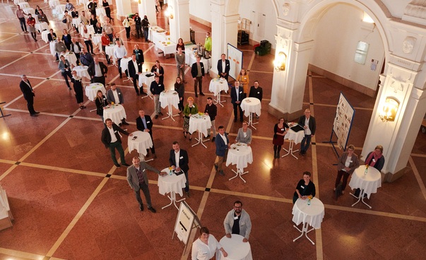 Gruppenbild unter Corona-Bedienungen. In einem großen Saal stehen jeweils zwei Personen links und rechts von einem Stehtisch.