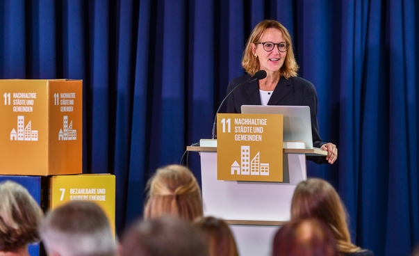 Sabine Drees vom Deutschen Städtetag steht am Rednerpult, das mit dem Symbol des globalen Nachhaltigkeitsziel Nr. 11 bestückt ist.