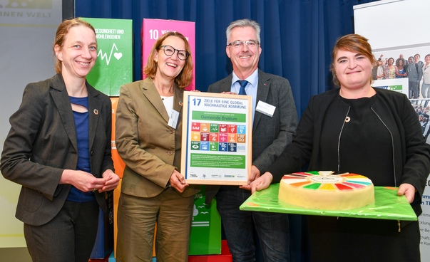 Drei Frauen und ein Mann im Halbporträt mit Urkunde und Torte in den Farben der Symbole der globalen Nachhaltigkeitsziele.