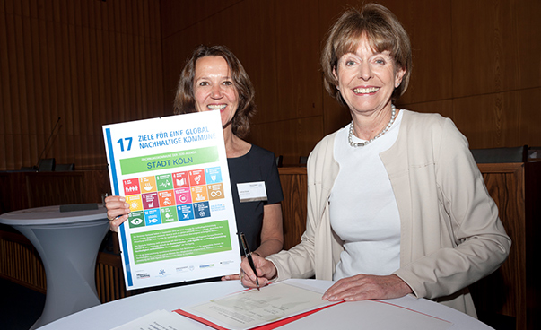 Die Kölner Oberbürgermeisterin Henriette Reker unterzeichnet die Musterresolution zur Agenda 2030. Sabine Drees vom Deutschen Städtetag hält die Anerkennungsurkunde in die Kamera.