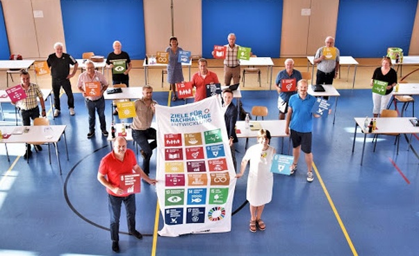in einer Turnhalle halten fünfzehn Frauen und Männer die Symbole der globalen Nachhaltigkeitsziele hoch.