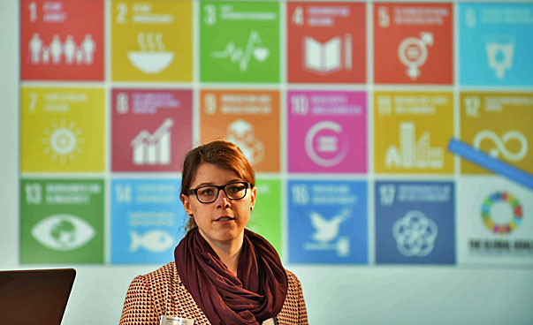 Eine Frau steht vor dem Hintergrund der Darstellung der nachhaltigen Entwicklungsziele und spricht in Richtung des Publikums. mehreren Personen vorstellt.
