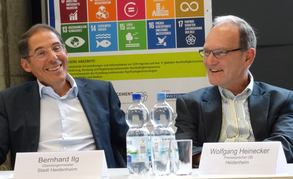 Zwei Männer sitzen vor einem Plakat, dass de Logos der globalen Nachhaltigkeitsziele zeigt.