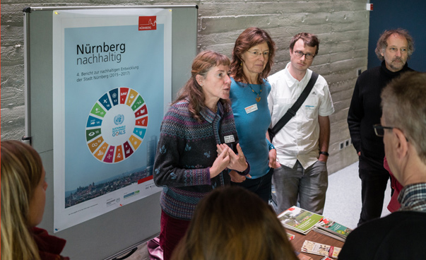 Eine Frau steht vor einem Plakat Nürnbergs mit den globalen Nachhaltigkeitssymbolen und spricht, während ihre andere Personen zuhören.