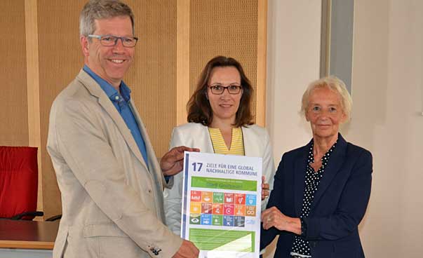 Drei Personen schauen in Richtung der Kamera, in der Mitte halten sie eine Urkunde mit den Symbolen der 17 nachhaltigen Entwicklungsziele. Foto: Pressestelle der Stadt Greifswald