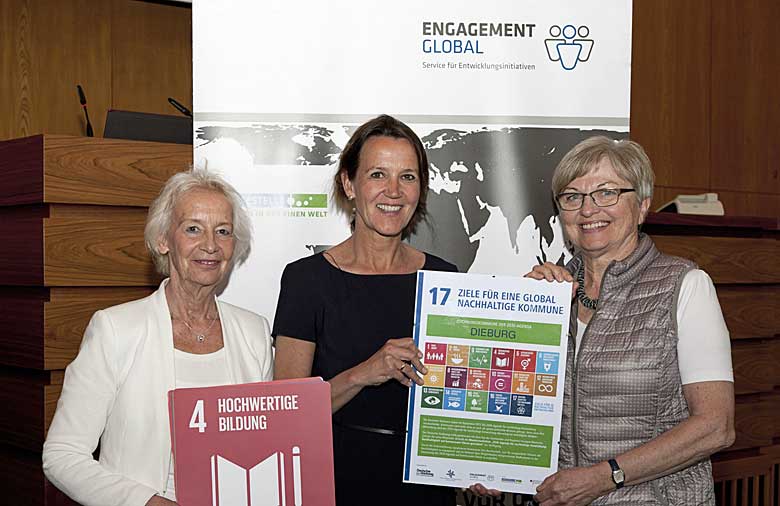 Drei freundlich lächelnde Personen halten eine Urkunde mit den Symbolen der 17 nachhaltigen Entwicklungsziele in Händen.