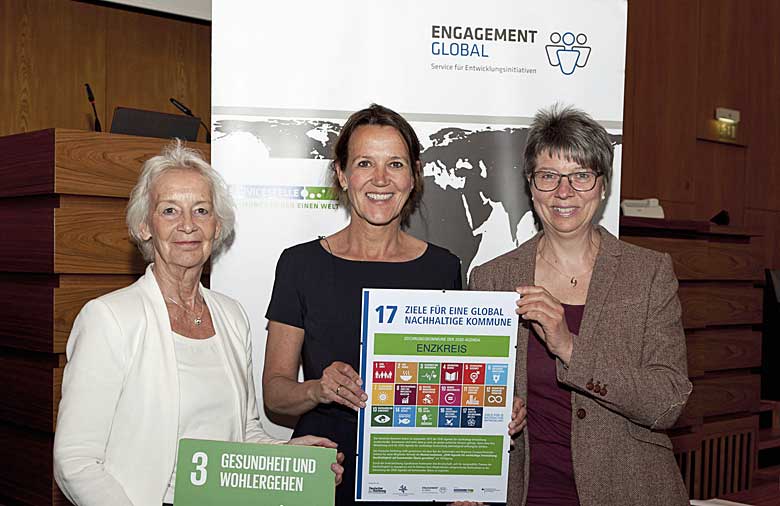 Drei freundlich lächelnde Personen halten eine Urkunde mit den Symbolen der 17 nachhaltigen Entwicklungsziele in Händen.