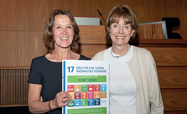 Zwei Frauen schauen freundlich in Richtung des Fotografen, sie halten eine Urkunde mit den Symbolen der 17 nachhaltigen Entwicklungsziele in Händen. Foto: Jo Hempel Photography