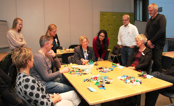 EIne Gruppe von circa 10 Personen sitzt oder steht um einen Tisch herum, auf dem Legobausteine liegen.