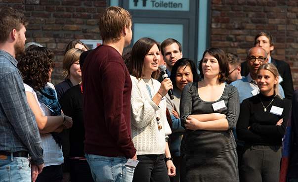 Eine junge Frau steht inmitten einer Gruppe Menschen und spricht ins Mikrofon. Ein junger Mann und eine weitere Frau stehen neben ihr und hören zu.