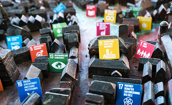Zwischen den Häusern einer metallenen Miniaturstadt stehen kleine Schilder mit den globalen Nachhaltigkeitssymbolen.