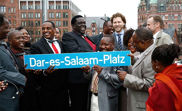Three men are laughing and holding a road sign with the inscription Dar es Salaam Square. In the background, brick buildings can be seen.