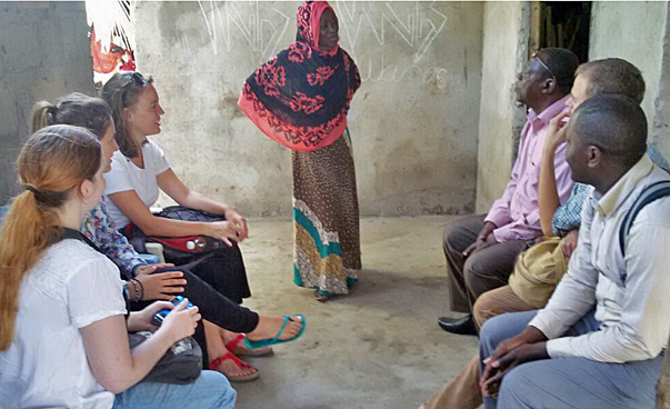 Eine Gruppe befindet sich in einem kleinen, einfachen Raum, in der Mitte steht eine Frau mit Kopftuch und spricht mit einer der Personen.