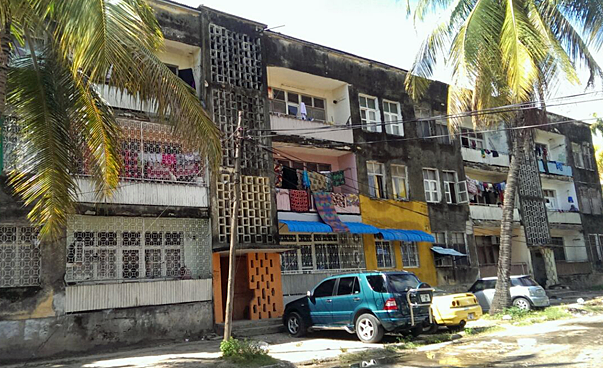 Vor einem mehrgeschossigen Gebäude mit Balkonen und Flachdach stehen Palmen.