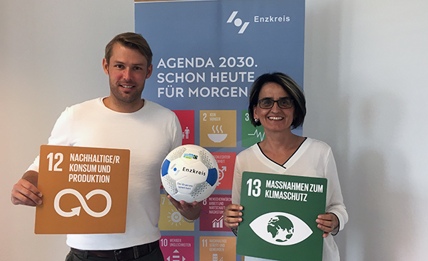 El Dr. Jannis Hoek y Edith Marques Berger sostienen ante la cámara tableros con los símbolos de sostenibilidad global número 12 y 13.