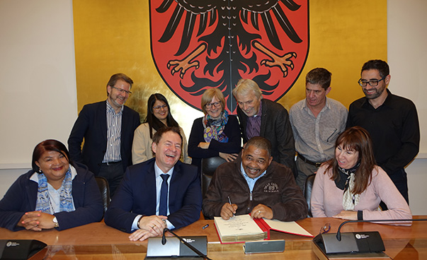 Alors que Conrad Poole signe le Livre d'or de la ville de Neumarkt, neuf autres personnes assises ou debout, dont le maire de Neumarkt, Thomas Thumann et Ralf Mützel, se réjouissent avec lui devant un grand blason coloré.