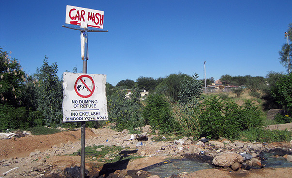 Un panneau portant l'inscription "Car Wash" est posé sur un sol rocailleux ; des buissons et des arbres sont visibles en arrière-plan.