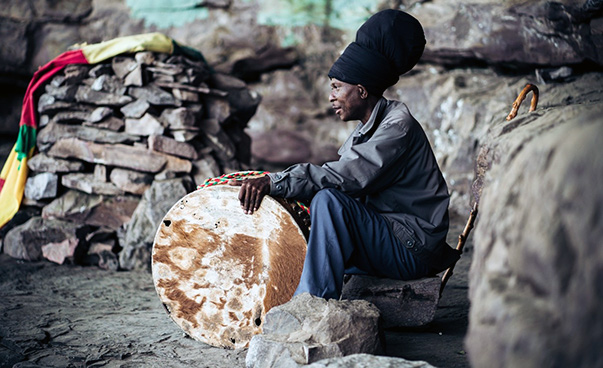 Eine Person mit einer sehr großen schwarzen Mütze hockt vor Felsen; in seiner Hand hält er eine Trommel.