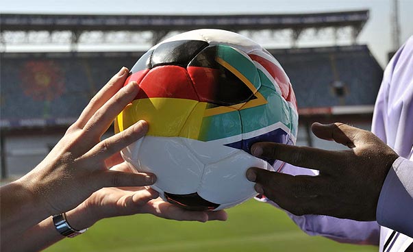 Vier Hände halten gemeinsam einen Fußball mit den Nationalfarben von Deutschland und Südafrika.
