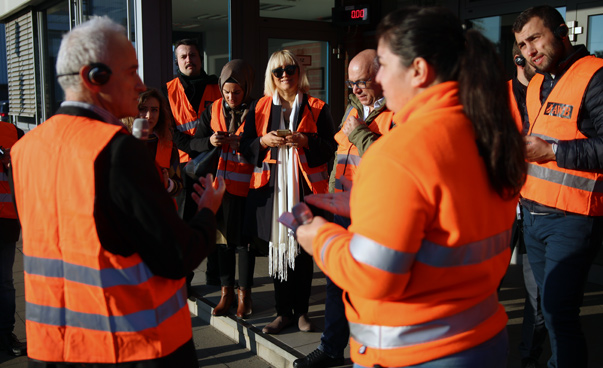 Ein Gruppe, mit orangen Warnwesten bekleidet, steht diskutierend vor einem Gebäude.