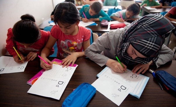 Three girls are sitting close together at a school desk and working on study materials. In the background, we can see other children in the classroom. Turkish municipalities are making efforts to create sufficient school places for Syrian children.
