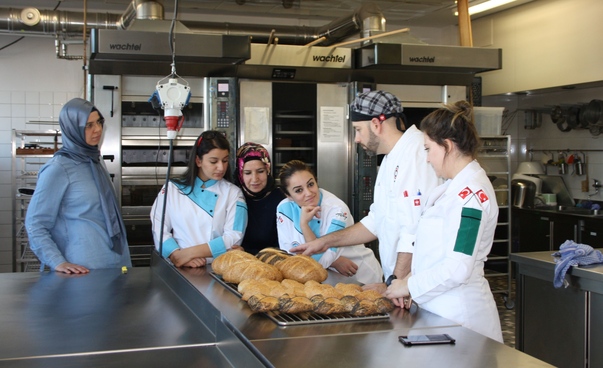 In einer professionellen Backstube schauen vier Frauen und ein Mann in weißer Berufsbekleidung auf ein mit Broten belegtes Backblech.