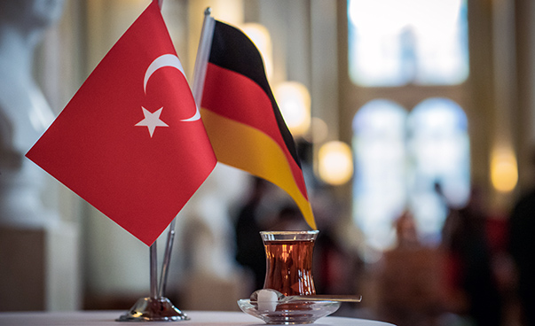 Ein deutsches und ein türkisches Fähnchen mit einem gemeinsamen Fuß stehen auf einem Tisch, rechts daneben steht ein türkischer Tee mit Zuckerwürfel.