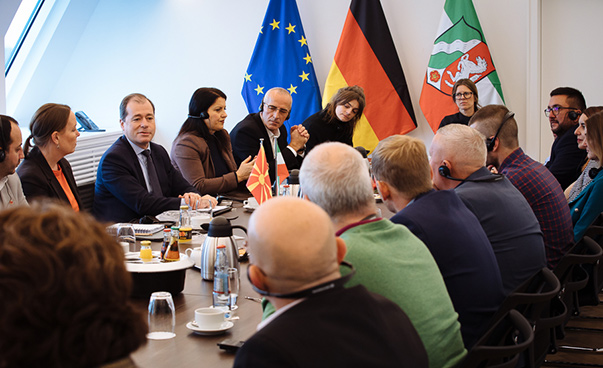 Rund ein Dutzend Personen sitzen an einem Tisch; im Hintergrund sind die europäische, die deutsche und die Flagge NRWs zu sehen.
