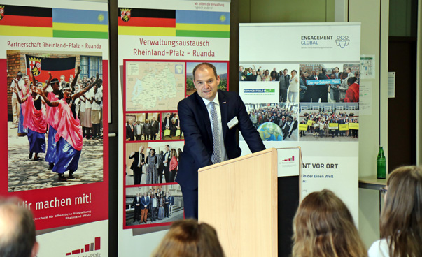A man stands at a lectern and speaks. Behind him are screens with pictures of the country partnership and of the Service Agency Communities in One World. In the foreground you can see the back heads of some people.