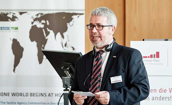 A man stands at a lectern and speaks, in the background is a world map to see.