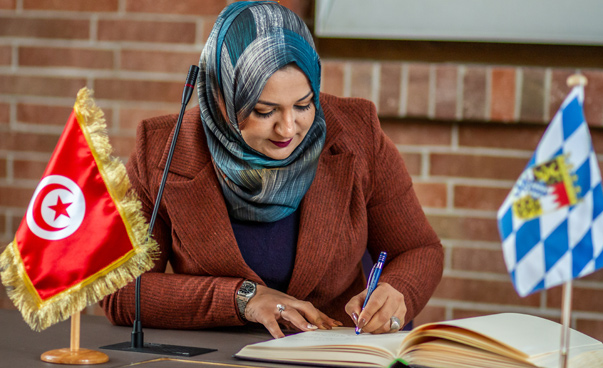 9)	Eine Frau trägt sich in ein Buch ein, vor ihr stehen eine tunesische und eine bayerische Tischflagge.