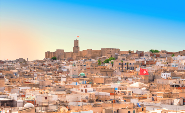 Vor strahlend blauem Himmel fällt der Blick auf eine arabische Altstadt; im Hintergrund ist eine alte Festungsanlage zu sehen.