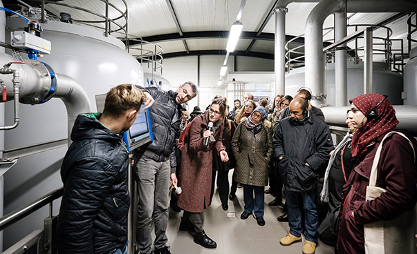 Un groupe d'une vingtaine de participants se tient en demi-cercle devant une usine d'une usine d'eau. Un homme montre un écran électronique et explique.