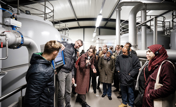 Eine Gruppe von circa zwanzig Teilnehmenden des Internationalen Netzwerktreffen Kommunaler Wissenstransfer Maghreb-Deutschland stehen in einem Halbkreis vor einer Anlage eines Wasserwerkes. Ein Mann zeigt auf einen elektronischen Bildschirm.