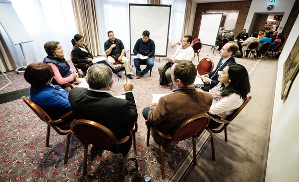 10 personas se sientan juntas en un círculo de sillas, discuten entre sí y toman notas.
