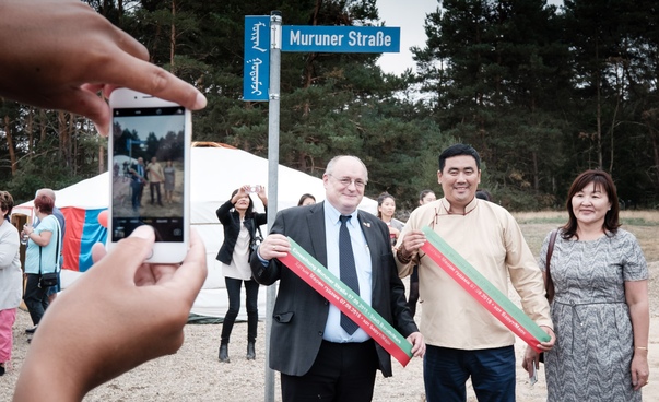 Zwei Männer und eine Frau stehen unter einem Straßenschild mit dem Ortsnamen einer mongolischen Kommune.