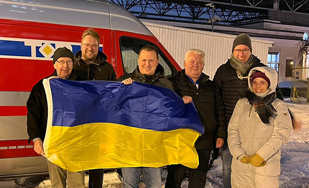 Six people stand in the snow in front of a lorry; they are holding a Ukrainian flag.
