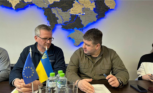 Two men sit at a table, talking; small Ukrainian and EU flags stand on the table.